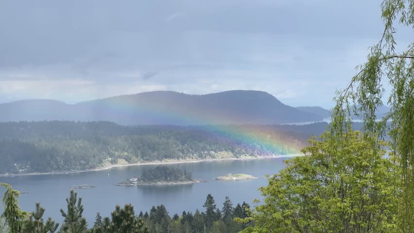 Vibrant rainbow arches over Saltspring Island’s forested coastline and rocky islets, viewed from a peaceful vacation home on a cloudy day.