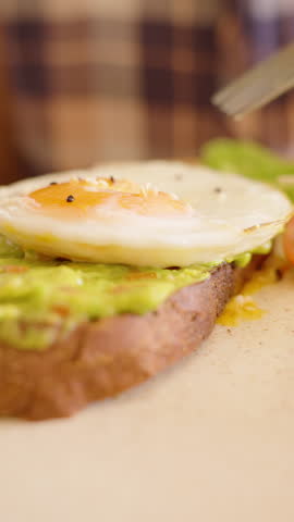 Close up of fork and knife cutting through runny yolk egg on avocado toast with salad on side in warm indoor setting, Healthy Eating Concept.