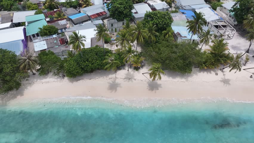 Aerial view of houses with colorful roofs nestled among lush green trees and palm trees along a pristine sandy beach, Maalhos, Baa Atoll, Maldives.