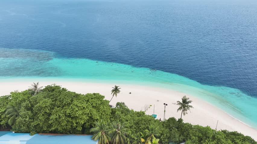 Aerial view of buildings nestled among lush trees, adjacent to a pristine beach with turquoise waters, creating a vivid contrast, Maalhos, Baa Atoll, Maldives.