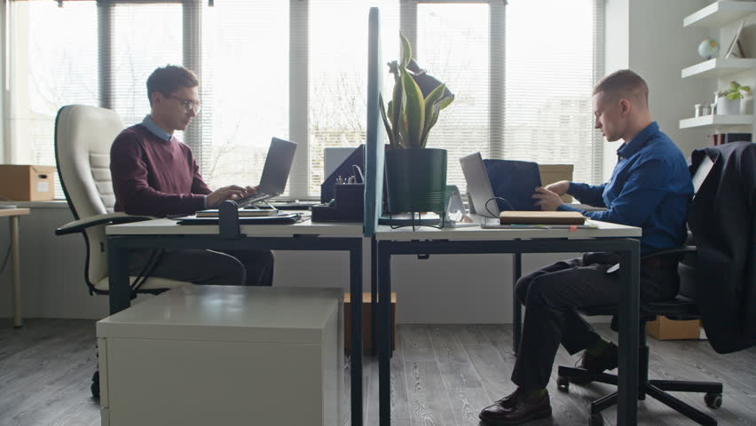 Side view of adult man handing folder with documents to male colleague sitting nearby at desk then returning to work on laptop in office workspace