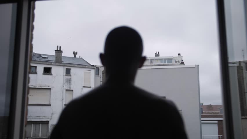 Silhouette man stands at window facing grey city rooftops on quiet overcast morning