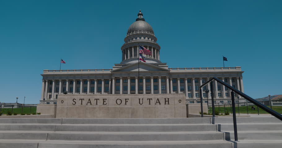 Front view of the Utah State Capitol building in Salt Lake City on a clear summer day, featuring the grand staircase, columns, dome, and flags against a bright blue sky.
