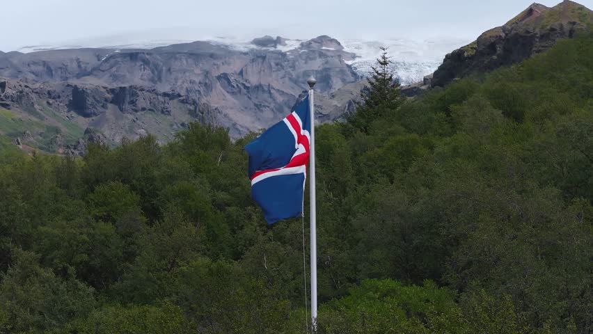 The Icelandic flag waves gently in a breeze, set against rugged cliffs, lush greenery, and rooftops, showcasing Iceland's dramatic landscapes.