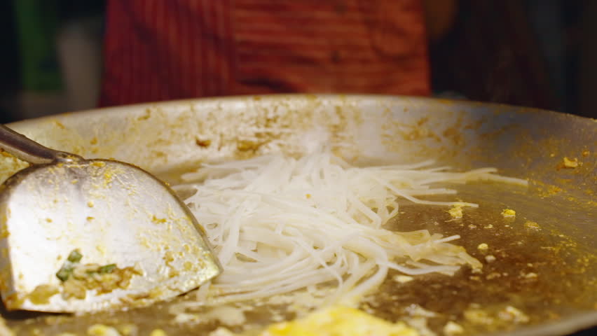 Close Angle Slow Motion Chef Pouring Sauce and Mixing Raw Pad Thai Noodles with Metal Utensils on Large Hot Pan, Street Food Concept.