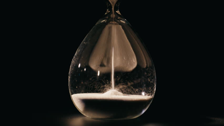 Elegant hourglass measuring minutes in dark room closeup. Macro view white sand falling inside glass clock counting seconds indoors. Transparent watch standing on black surface capturing time flowing.