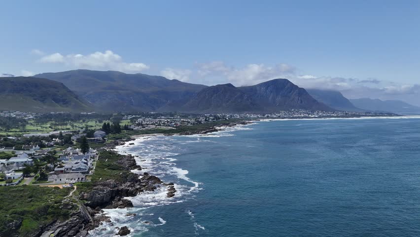 4K drone footage of coastal cliffs and ocean waves crashing against rocks in Hermanus, near Cape Town, South Africa. Scenic shoreline with mountain backdrop along the South Atlantic coast.