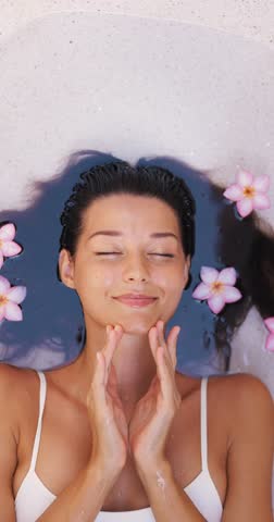 Close-up vertical slow motion of a woman relaxing in clear water surrounded by pink plumeria flowers, gently caressing her face. Ideal for skincare, wellness, beauty and spa promos.