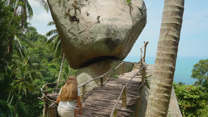 Woman Walking on Narrow Wooden Bridge Toward Huge Balanced Rock With Ocean View in Background From Elevated Angle at Overlap Stone Samui, Exploration Adventure Concept.