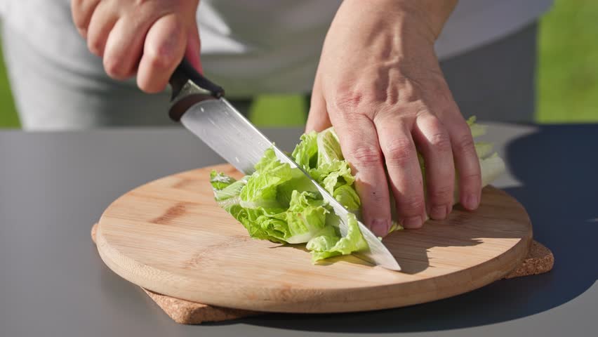 chef cuts fresh green romaine lettuce leaves on cutting board to prepare a healthy salad. outdoor cooking, food closeup