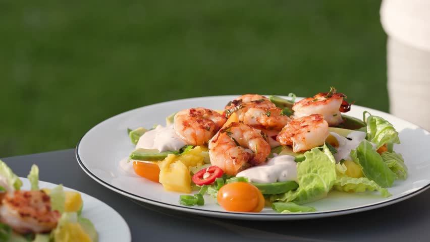 Chef shows portion of freshly prepared healthy vegetable salad with grilled shrimps on plate. outdoor cooking, food close-up