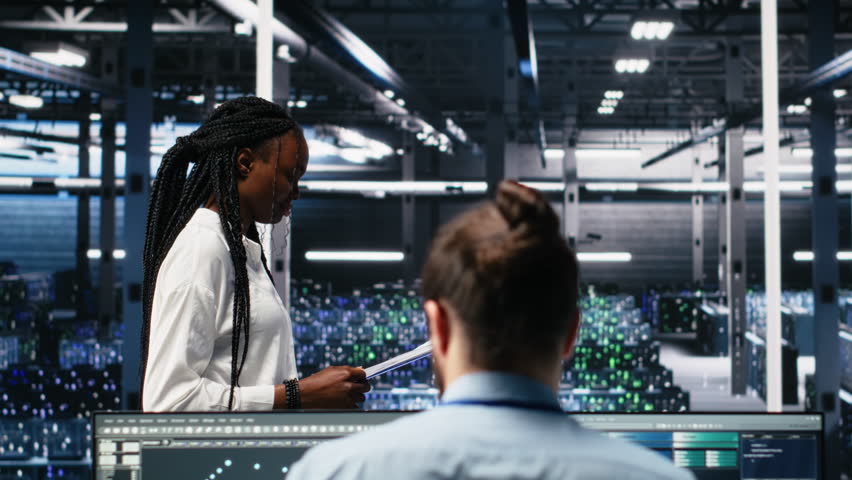 Data center engineers managing AI systems for uptime and efficiency, looking over paperwork files. African american woman and Asian man reading documents in multiethnic server hub, camera A