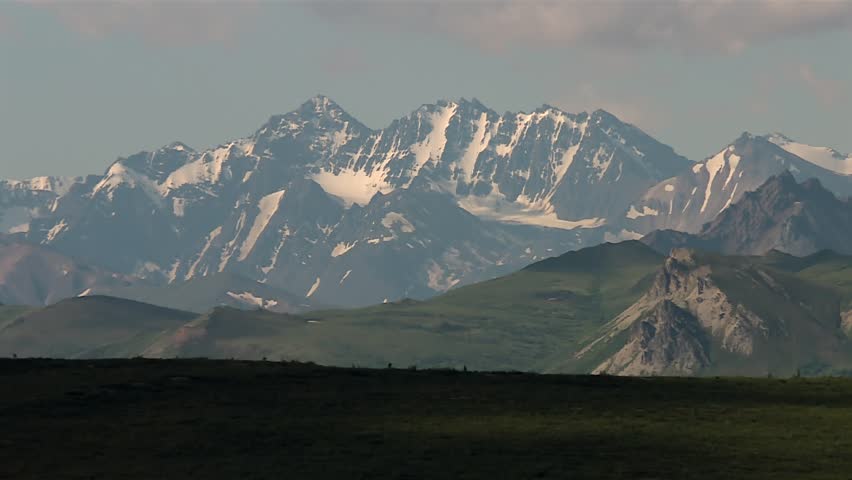 Snow covered peaks with rugged rocky ridges and green foothills in Denali National Park, Alaska. Static daytime view.
