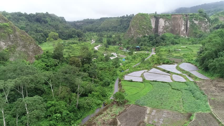 Drone zoom in over lush valley in West Sumatra, Indonesia, revealing hidden landscape behind soft clouds. Peaceful tropical scenery. and a car drive by surround by trees and rice paddy