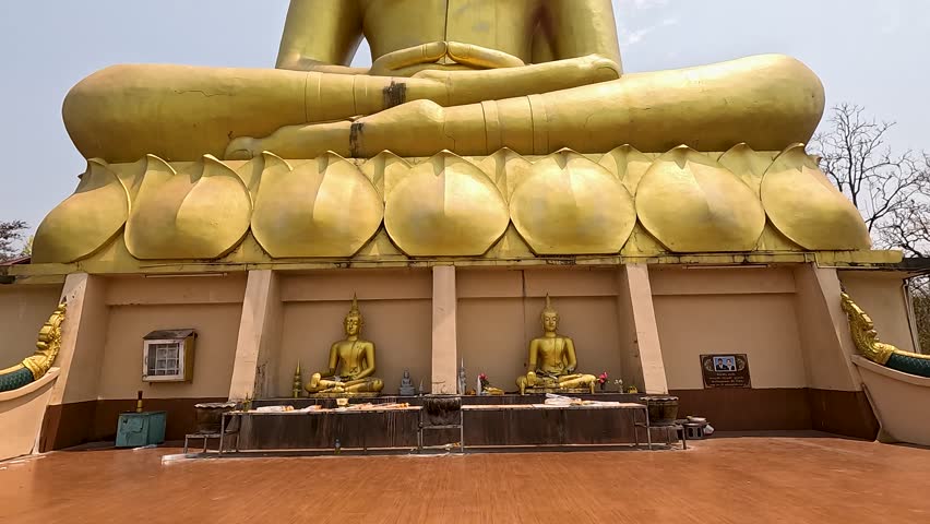 Camera tilts upward from base to head of a massive golden Buddha statue at an outdoor temple, under bright daylight and clear sky