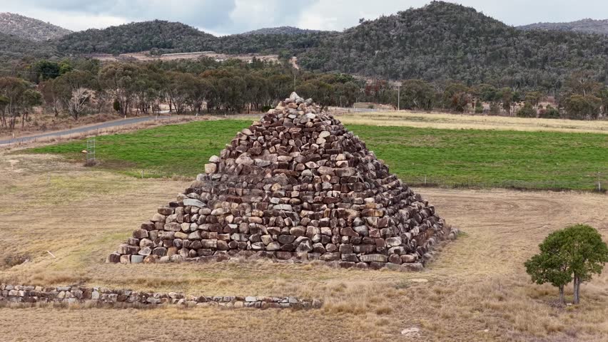 Drone camera smoothly circles a large granite rock pyramid in a dry, grassy field near Ballandean, Queensland, under natural daylight with steady movement