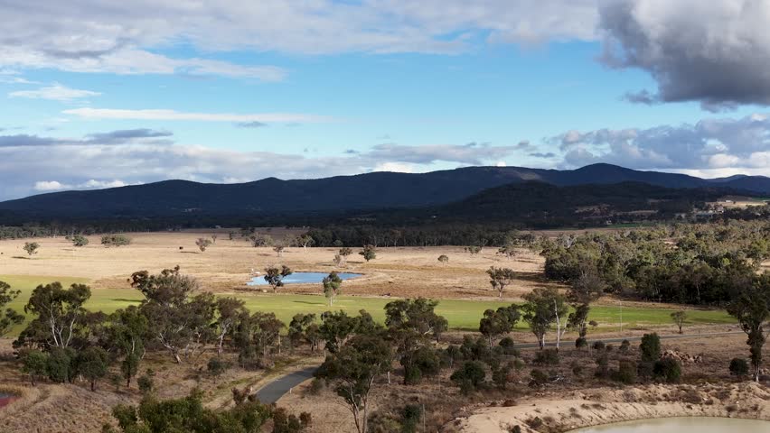Aerial footage pans across a rural Australian landscape with farmland, pond, scattered trees, and distant hills under partly cloudy daylight