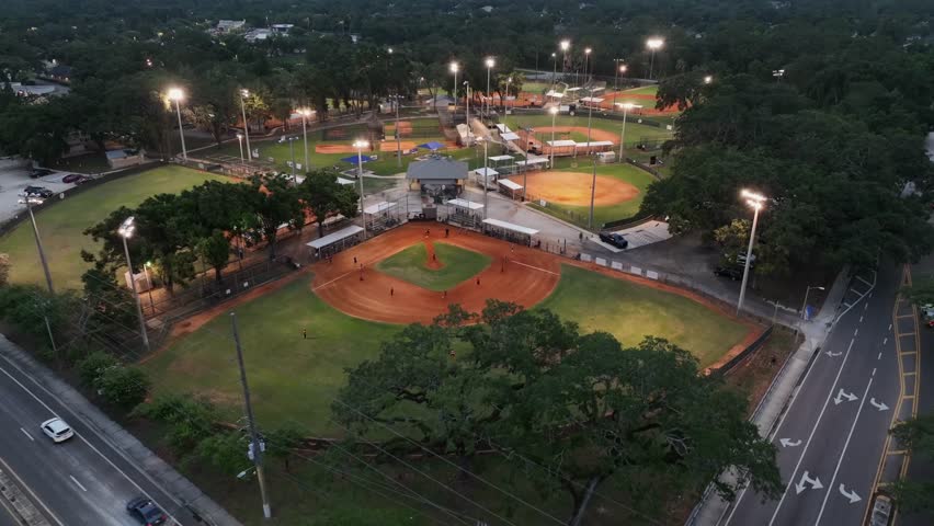 Several baseball fields and playing people in the evening with lighting floodlight. Aerial flyover shot. Florida suburb district in summer. Sport area with several courts.