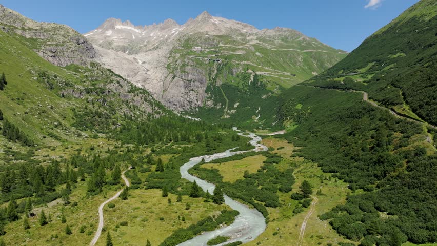 Establishing shot of alpine river, green valley leading toward rugged Furkapass