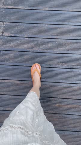 POV. First - person view of walking on a wooden boardwalk, showing lower body in a white lace skirt and yellow flip - flops. Casual, daily life outdoor scene.