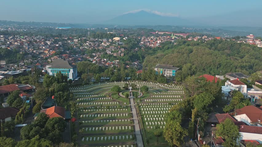Semarang, Indonesia - July 16 2025 : High-angle aerial shot over a serene war cemetery in Indonesia. The view expands to a sprawling city below and a majestic mountain on the horizon.