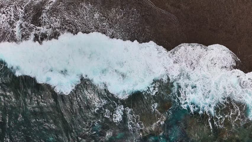 Aerial view of ocean waves crashing onto a rocky shoreline, with frothy white surf meeting dark coastal rocks and clear blue water forming turbulent patterns as it flows toward the beach