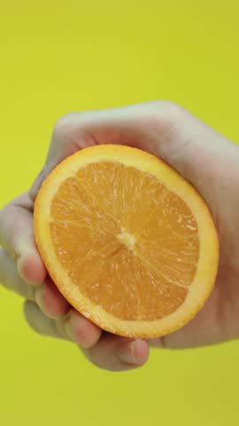 Orange being squeezed by hand with juice splashing against yellow background, close-up slow motion