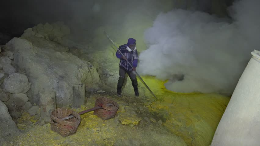 Wide shot shows a masked miner in Kawah Ijen crater using a long pole to break cooling sulfur near a steaming vent, with traditional woven baskets ready for the heavy load.