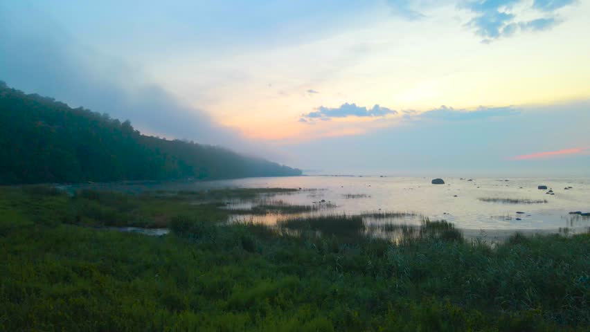 Stunning aerial drone footage gliding over grassy and reed filled beach area and continiues to fly over Baltic ocea sea during orange sunset, steep cliff bank in fog and mist visible on the left.
