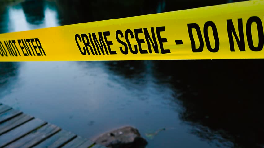 Close up video of a yellow colored Crime Scene Do Not Enter police caution ribbon tape pulled in front of a dark blue lake that is reflecting trees during a rainy evening. Wooden deck at the bottom.