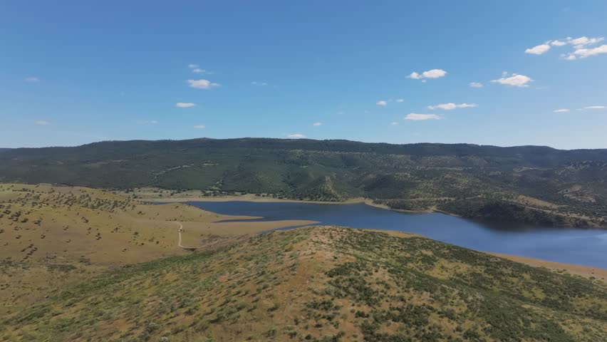 Low forward flight over golden hills and green shrubs around the Cijara reservoir, full from spring rains. Blue-green water reflects sky and forests under a sunny May sky in Badajoz, Spain.