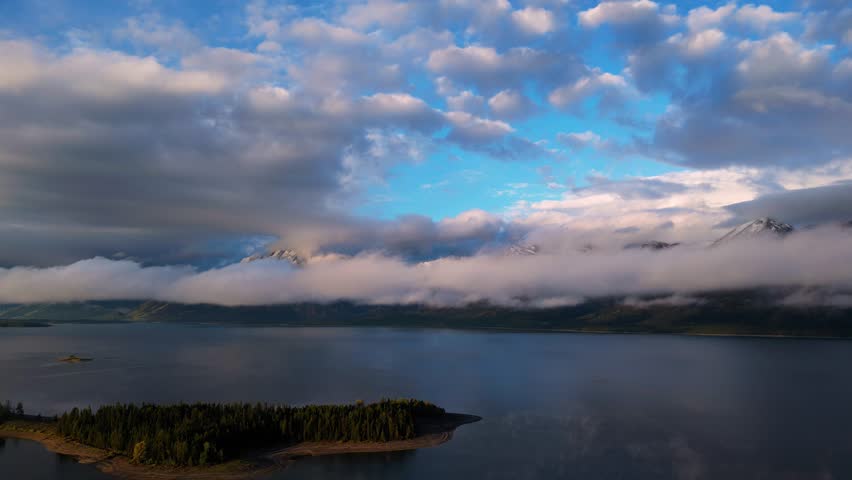 Aerial view of the serene Jackson Lake reflecting the sky, surrounded by evergreen forests and snow-capped mountains peeking through the clouds, Wyoming, United States.
