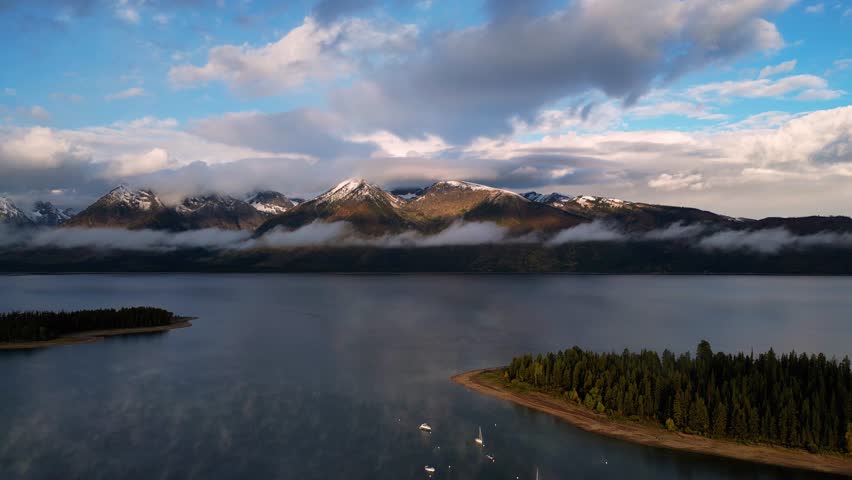 Aerial view of majestic, snow-capped mountains shrouded in ethereal clouds reflected in the tranquil lake dotted with boats, Teton County, United States.