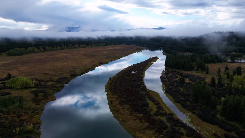 Aerial view of a winding river reflecting the sky, cutting through a landscape of brown fields and green forests under a cloudy sky, Teton County, United States.