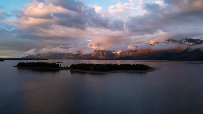 Aerial view of Jackson Lake reflecting the sky, with mountains partially shrouded in clouds, exhibiting a blend of warm and cool tones, Teton County, United States.