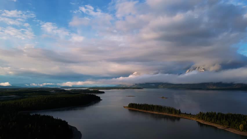 Aerial view of an island covered with trees in a lake, with snow-capped mountains partially obscured by clouds, Teton County, United States.