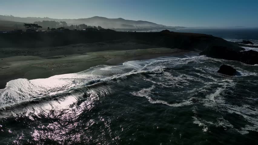 Aerial view of powerful waves crashing onto a dark sandy beach contrasting with the rugged cliffs and hazy horizon, California, United States.