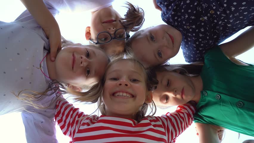 Low angle view of happy little children in circle look at camera embrace and laugh in summer park. Team kids play outdoors. Close up of face girls and boys