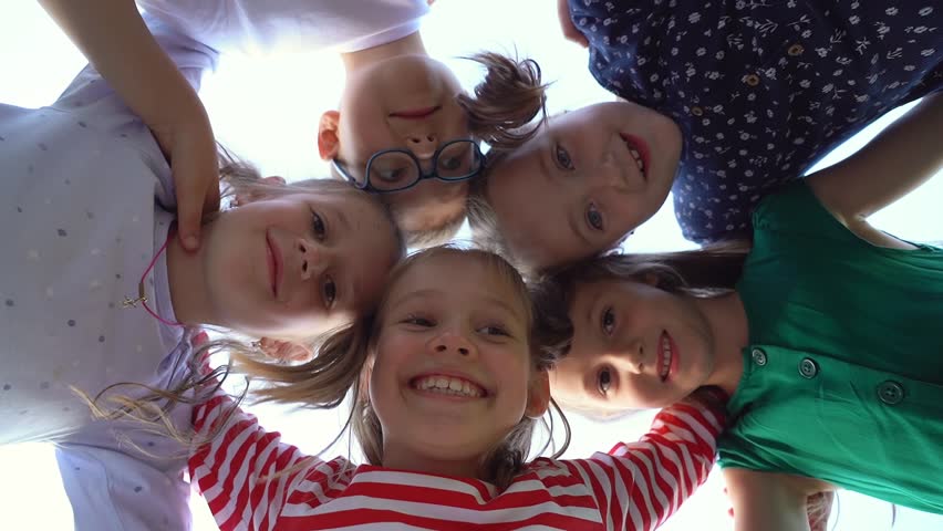 Low angle view of happy little children in circle look at camera embrace and laugh in summer park. Team kids play outdoors. Close up of face girls and boys