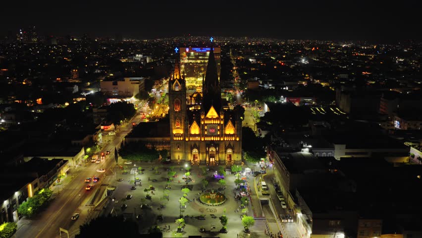 Drone circles Templo Expiatorio at night highlighting intricate Gothic features and colorful stained glass from above, establishing orbit over park in Guadalajara
