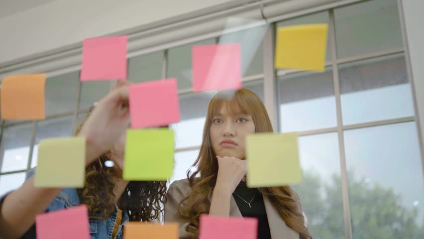 Asian creative business women are writing note on the text sheet after thinking new idea and planning project with colleague, Two employee brainstorming idea on glass wall and sticky