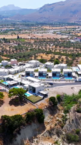 Vertical drone shot orbiting upscale holiday homes on a hill, in sunny Greece