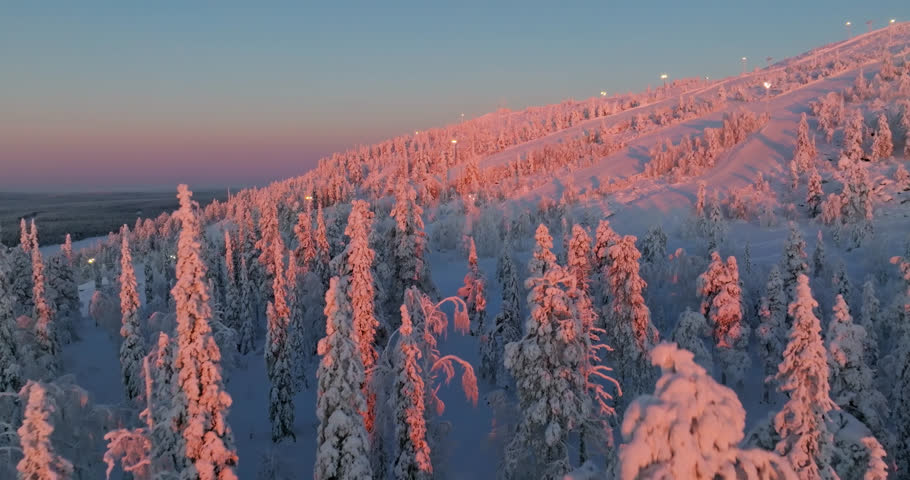 Drone flying over glowing forest and ski slopes in Lapland, winter sunrise