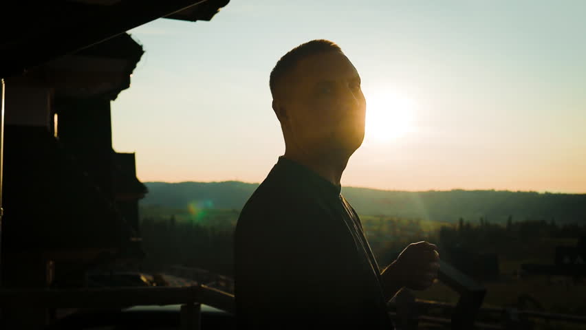 A man stands on a balcony looking out at a peaceful sunrise over a rural landscape. His silhouette is softly lit by morning light, evoking a sense of calm, hope, and new beginnings. Ideal for themes