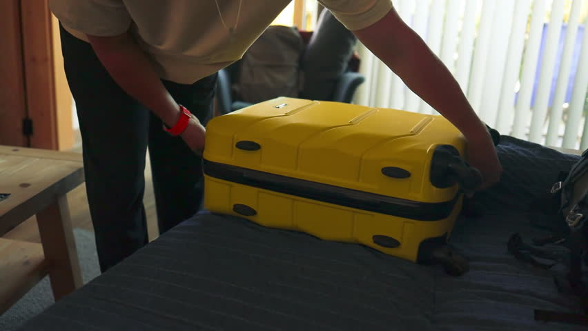 A man is unpacking a yellow suitcase in a cozy room, taking out travel clothes and essentials. Soft natural light shines through the blinds, creating a calm and homey atmosphere after returning from a