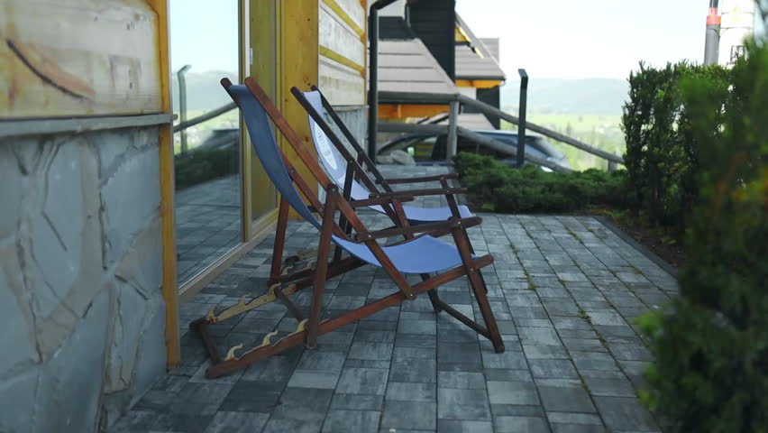 Two empty wooden deck chairs rest on a stone terrace outside a rustic house, surrounded by greenery and overlooking a peaceful mountain landscape. A tranquil vacation setting perfect for relaxation