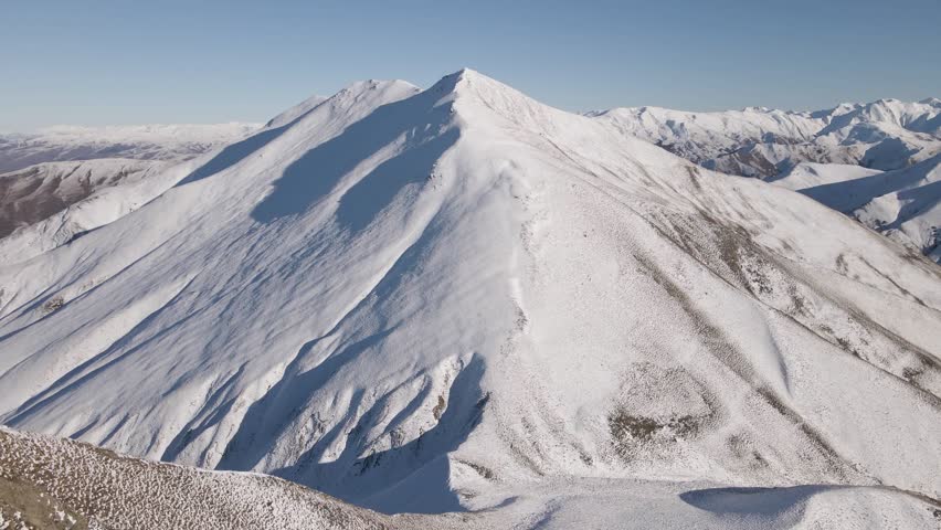 Aerial shot flying toward a snow-covered mountain near Lindis Pass while slowly tilting up to reveal the steep ridgeline and surrounding alpine landscape