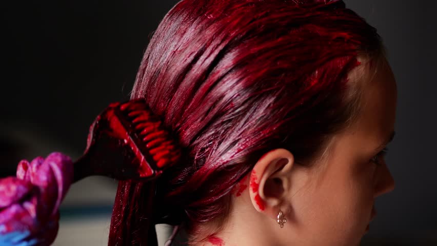 A close-up side view of a person's hair being dyed a vibrant red, with a gloved hand applying the color using a brush.