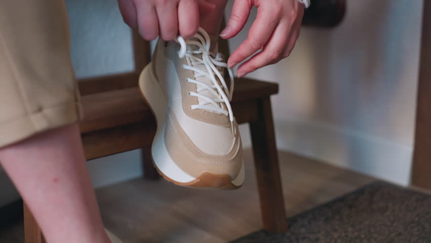 Closeup leg view of guest untying laces to remove beige canvas shoe while seated on wooden bench, preparing for indoor session in calm setting with warm lighting and cozy room decor details around