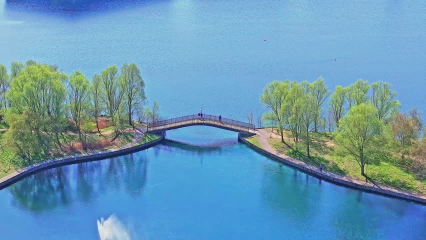 Ascending slow aerial view of Doncaster Lakeside showing pedestrian bridge, young green trees, clear blue water, and shoreline reflections under bright spring sunlight.
